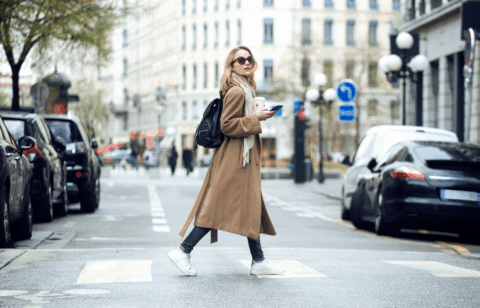 A woman wearing a long coat and backpack crosses a city street while holding a smartphone and a coffee.