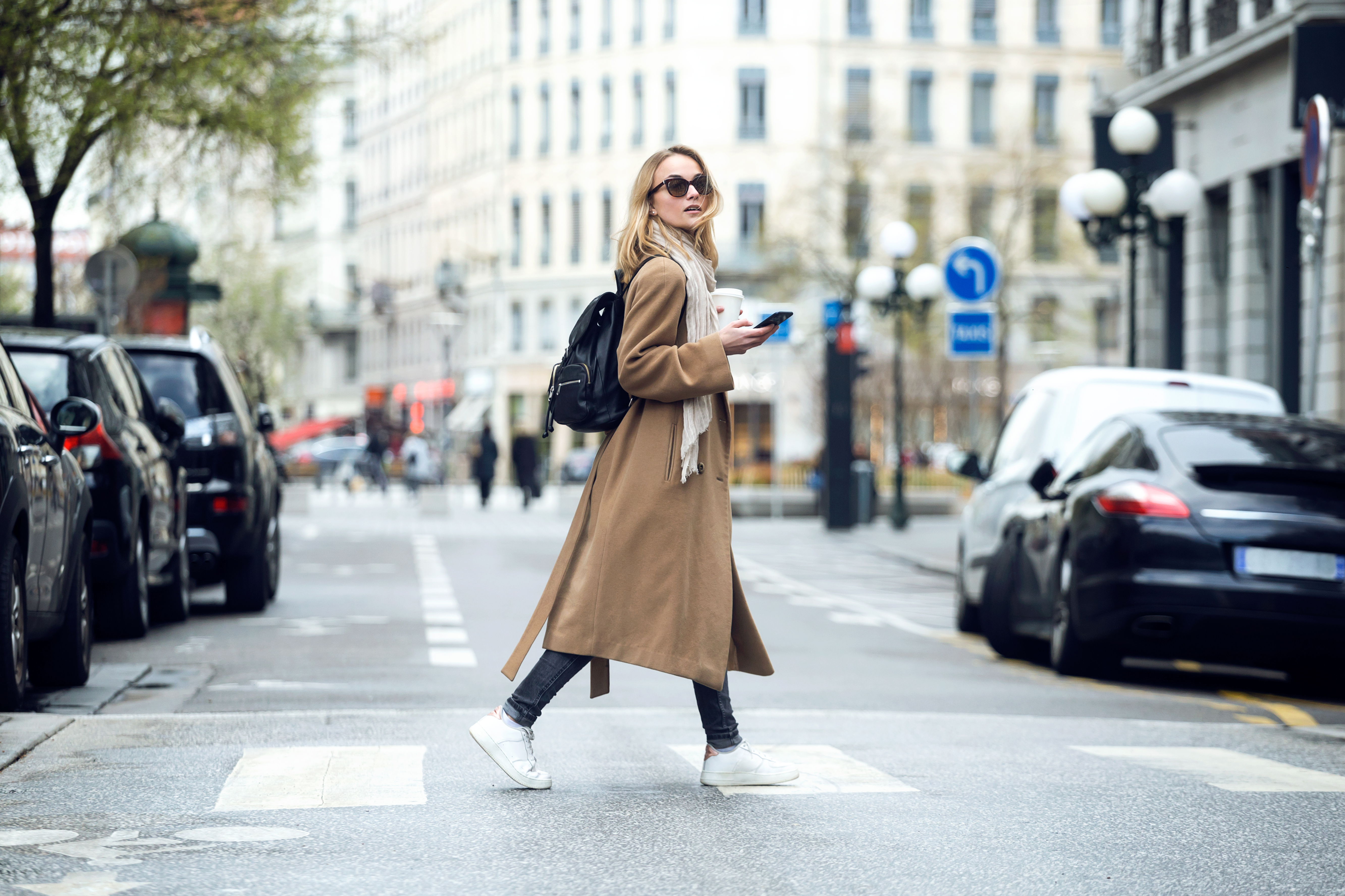 A woman wearing a long coat and backpack crosses a city street while holding a smartphone and a coffee.