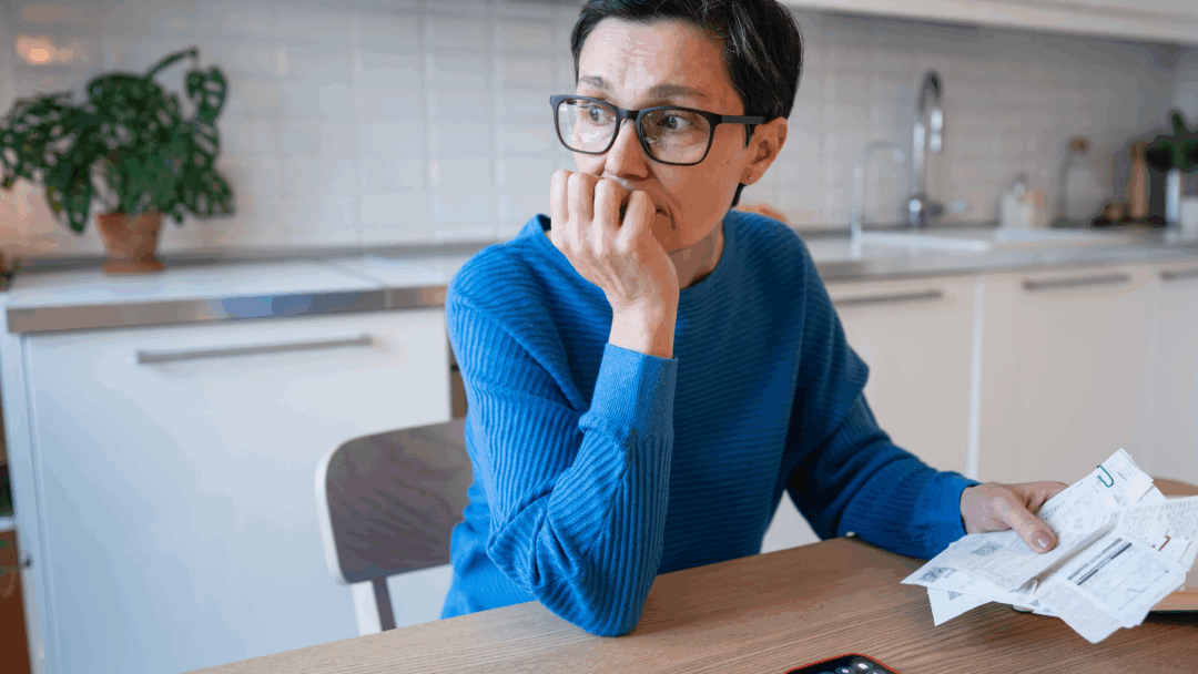 Person sitting at a kitchen table holding bills and looking off to the side with a thoughtful expression.