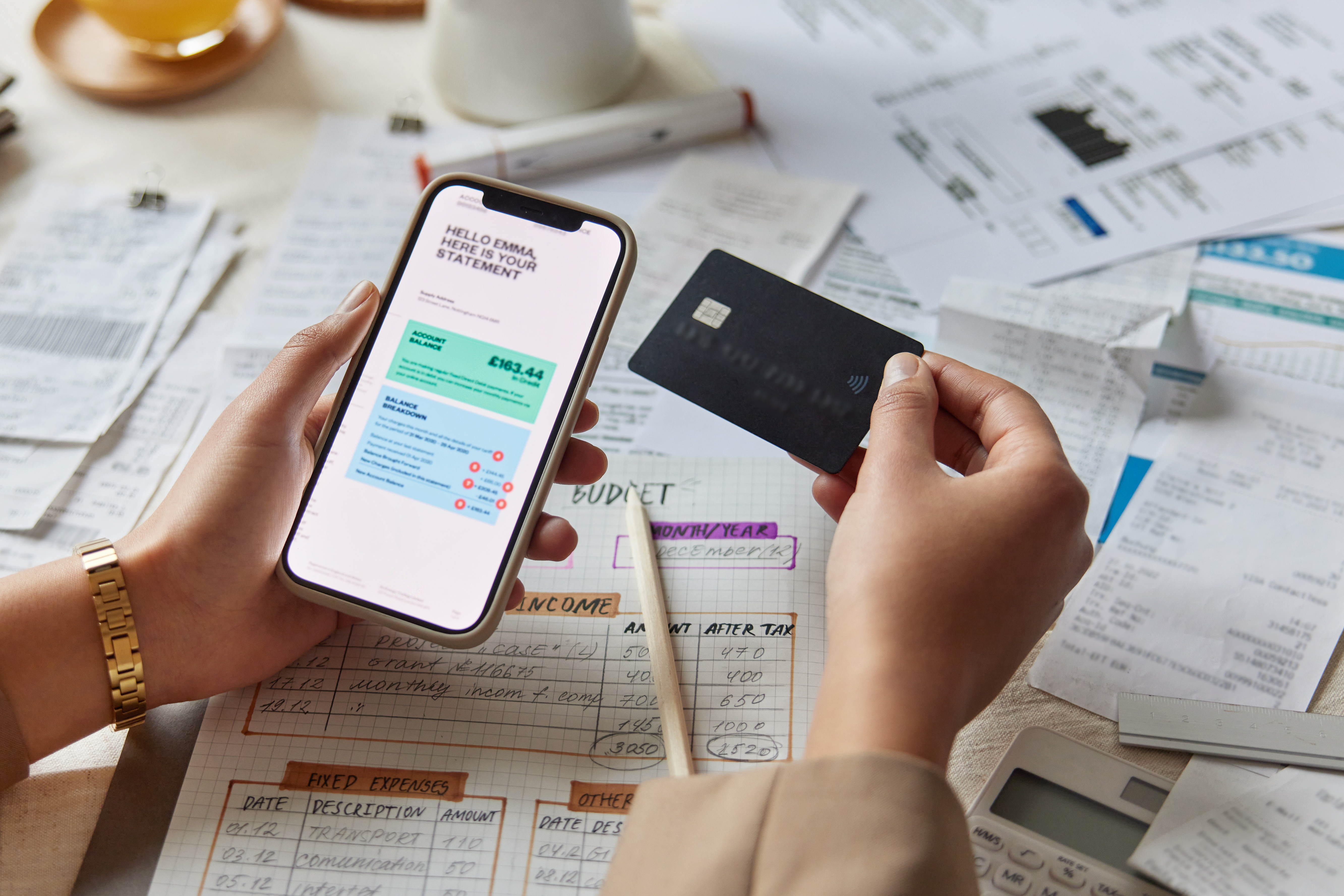 Hands hold a smartphone showing a bank statement and a credit card above a table covered with bills, receipts, and a handwritten budget.