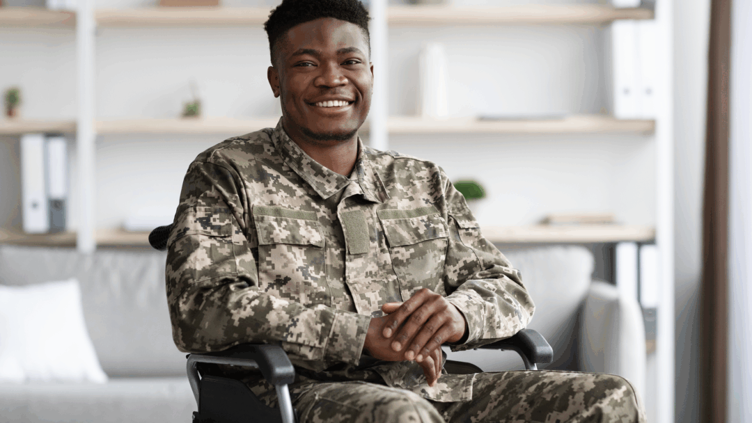 Veteran in camouflage uniform seated in a wheelchair, smiling at the camera.
