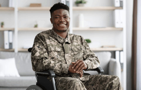 Veteran in camouflage uniform seated in a wheelchair, smiling at the camera.