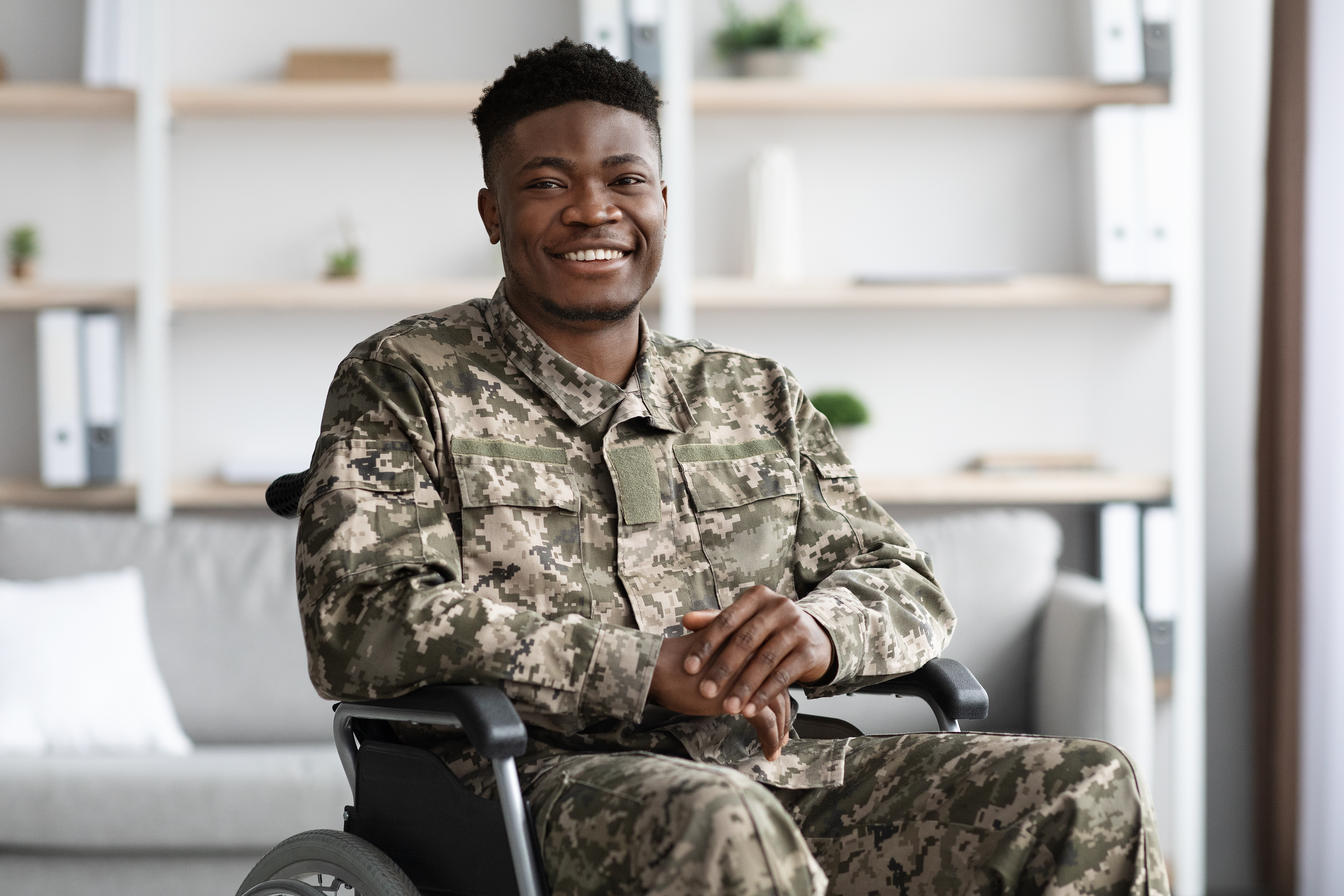 Veteran in camouflage uniform seated in a wheelchair, smiling at the camera.