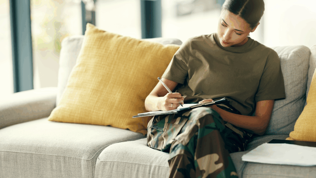 A service member sits on a couch and writes on a clipboard while reviewing paperwork.