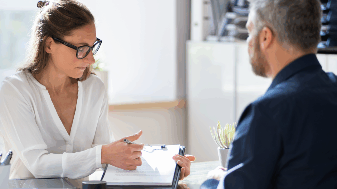 A woman wearing glasses points to a document on a clipboard while speaking with a man seated across a desk.