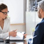 A woman wearing glasses points to a document on a clipboard while speaking with a man seated across a desk.