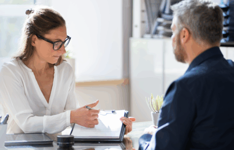 A woman wearing glasses points to a document on a clipboard while speaking with a man seated across a desk.