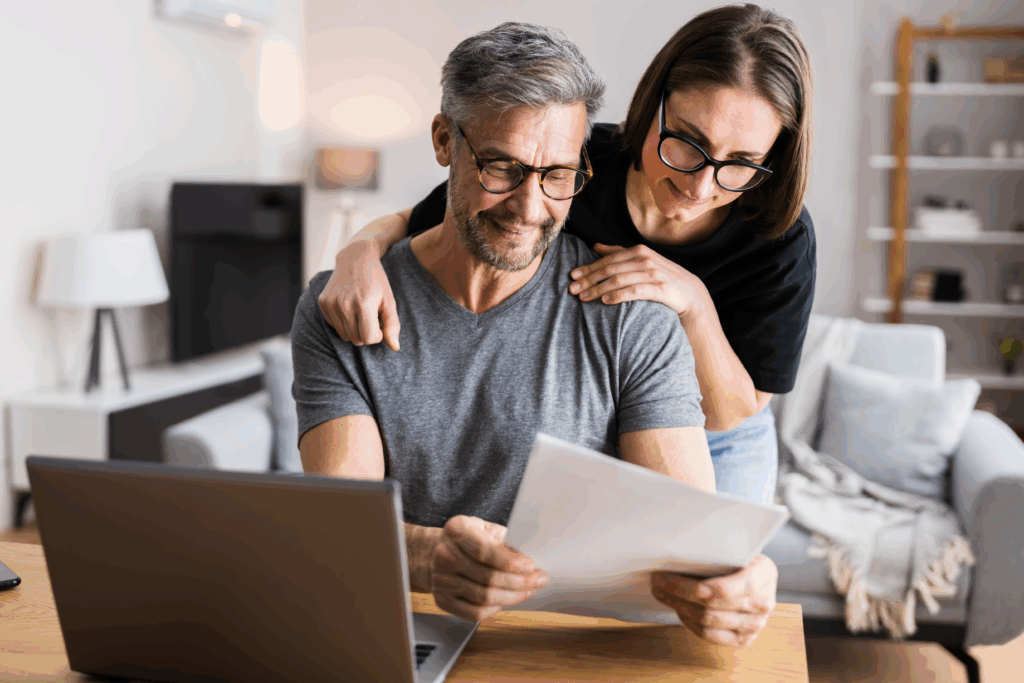 A middle-aged couple reviews paperwork together at a table while looking at a laptop in their living room.