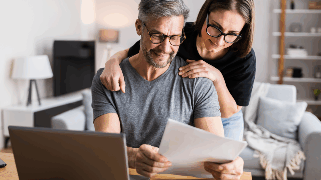 A middle-aged couple reviews paperwork together at a table while looking at a laptop in their living room.