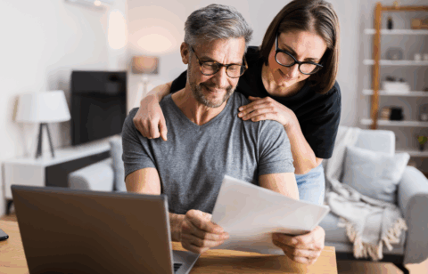 A middle-aged couple reviews paperwork together at a table while looking at a laptop in their living room.