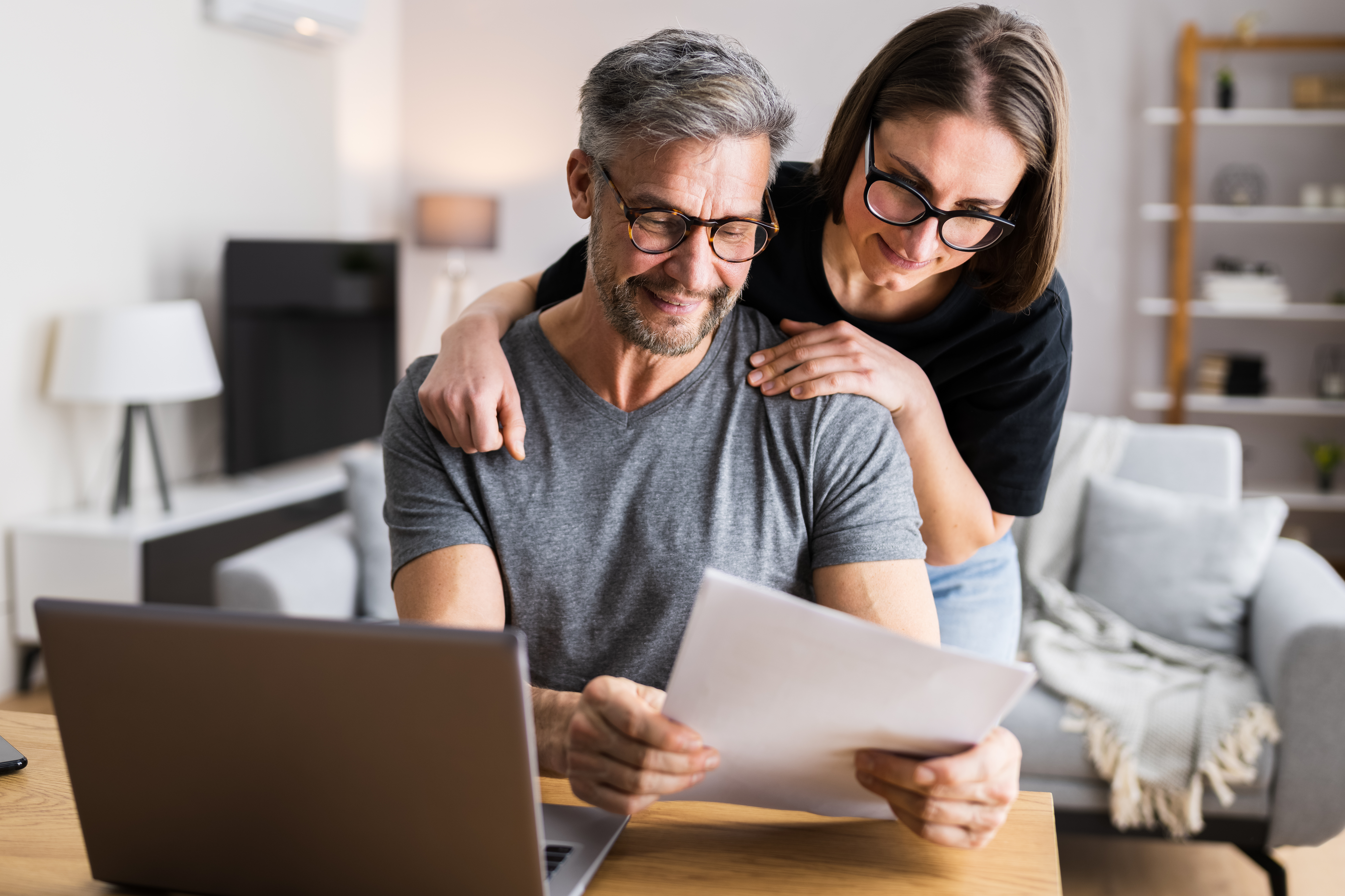A middle-aged couple reviews paperwork together at a table while looking at a laptop in their living room.