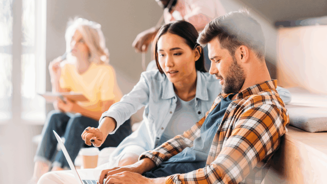 A woman points at a laptop screen while working with a man during a group study or workshop.