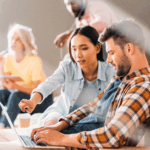 A woman points at a laptop screen while working with a man during a group study or workshop.