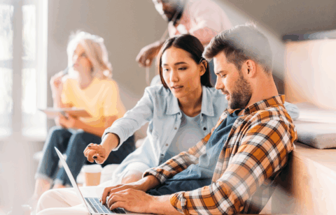 A woman points at a laptop screen while working with a man during a group study or workshop.