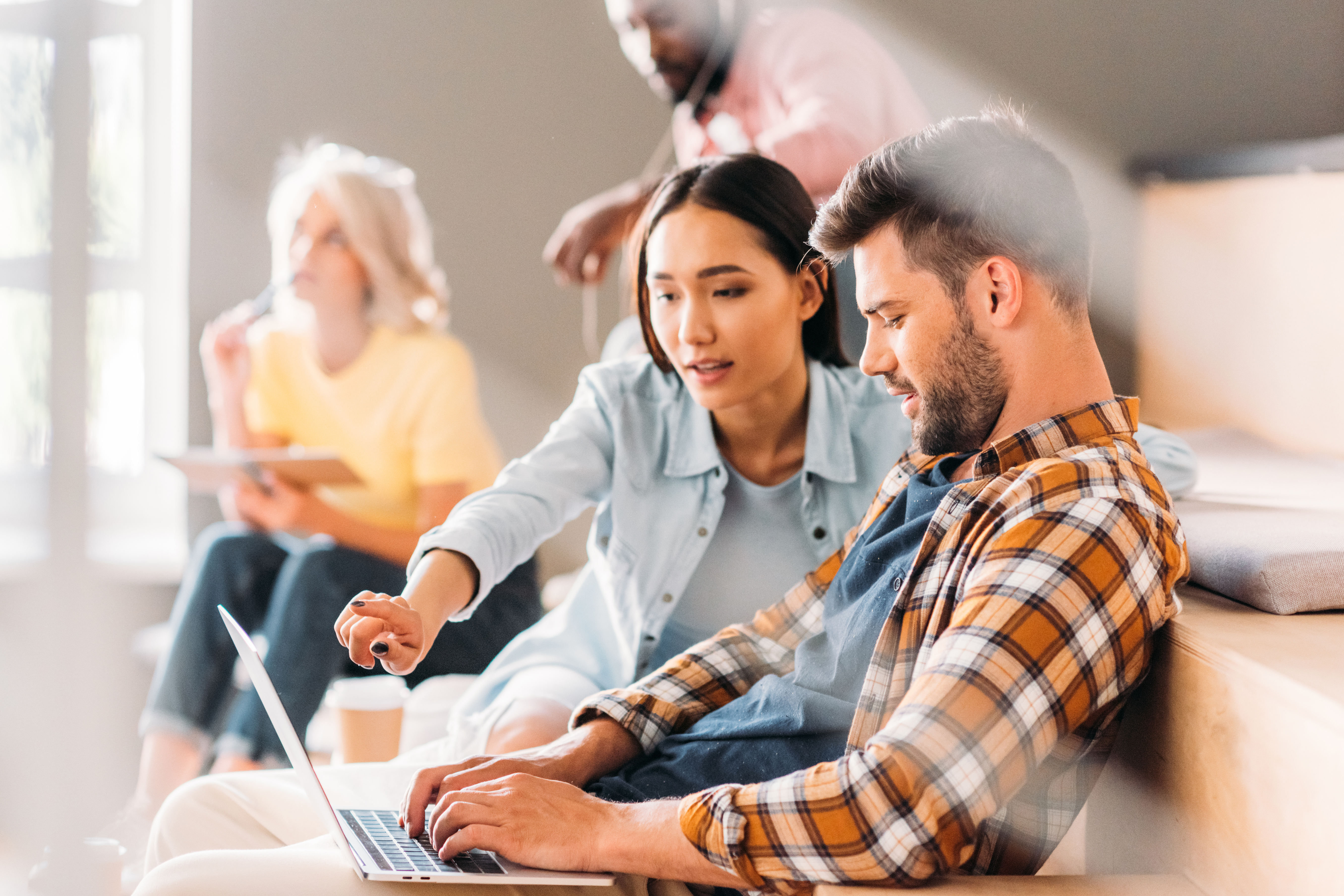 A woman points at a laptop screen while working with a man during a group study or workshop.