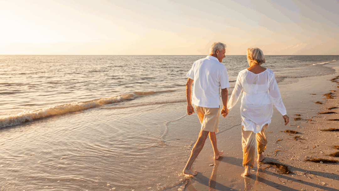 An older couple walks hand in hand along the beach at sunset.