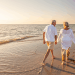 An older couple walks hand in hand along the beach at sunset.