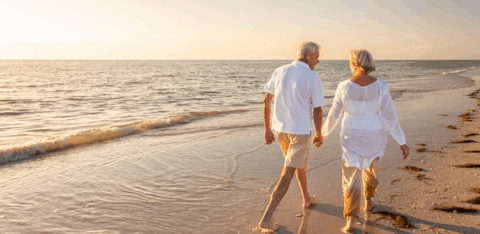 An older couple walks hand in hand along the beach at sunset.