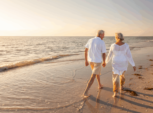 An older couple walks hand in hand along the beach at sunset.