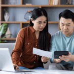 A couple sits on a couch reviewing a bill together while using a calculator and a laptop.