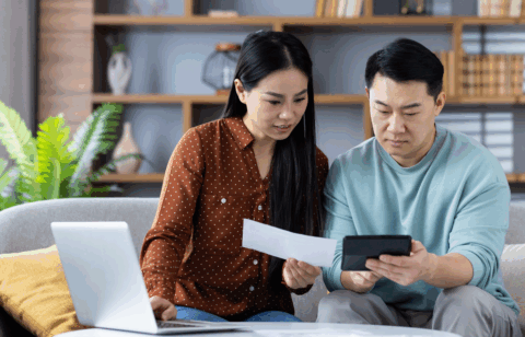 A couple sits on a couch reviewing a bill together while using a calculator and a laptop.
