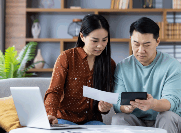 A couple sits on a couch reviewing a bill together while using a calculator and a laptop.