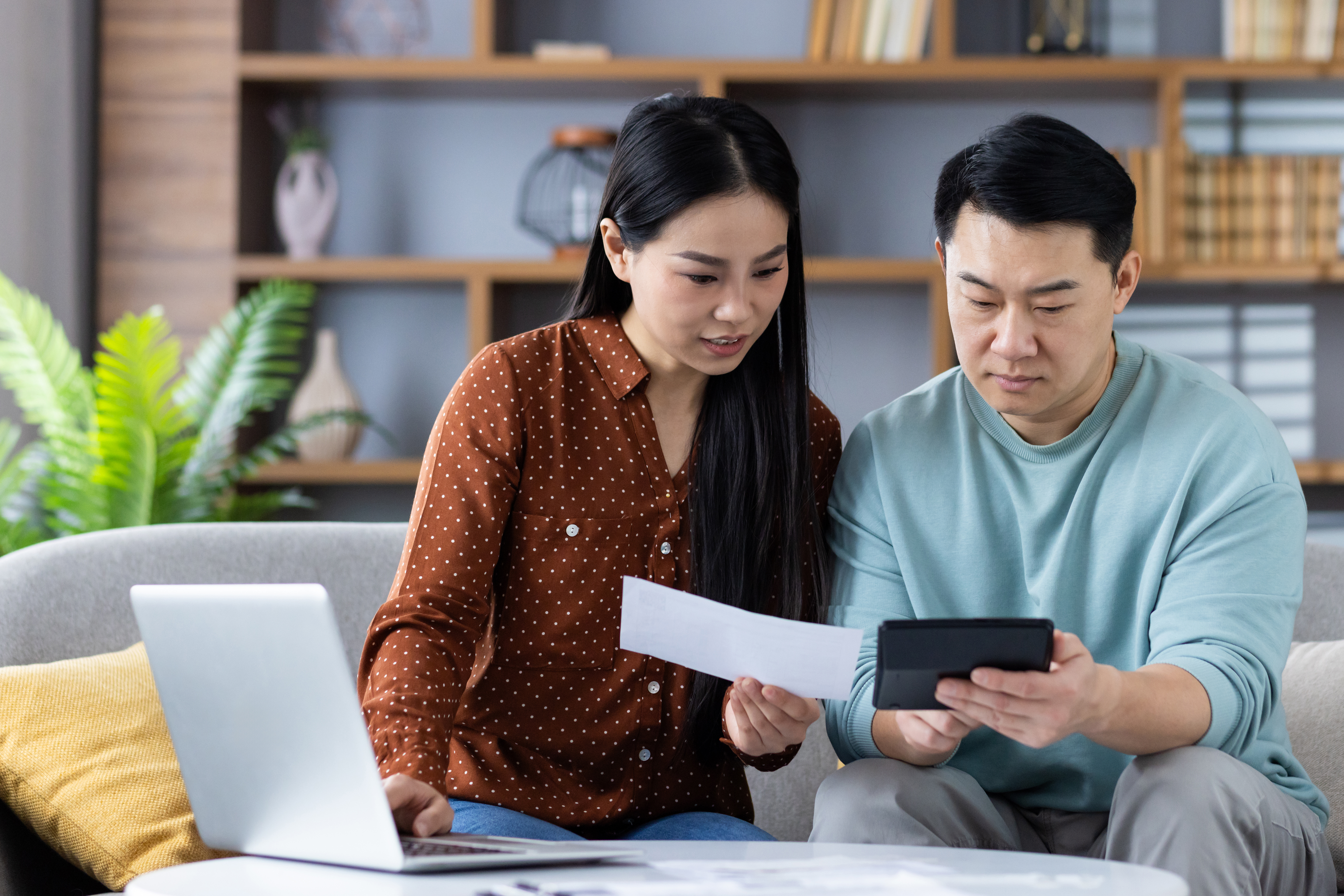 A couple sits on a couch reviewing a bill together while using a calculator and a laptop.