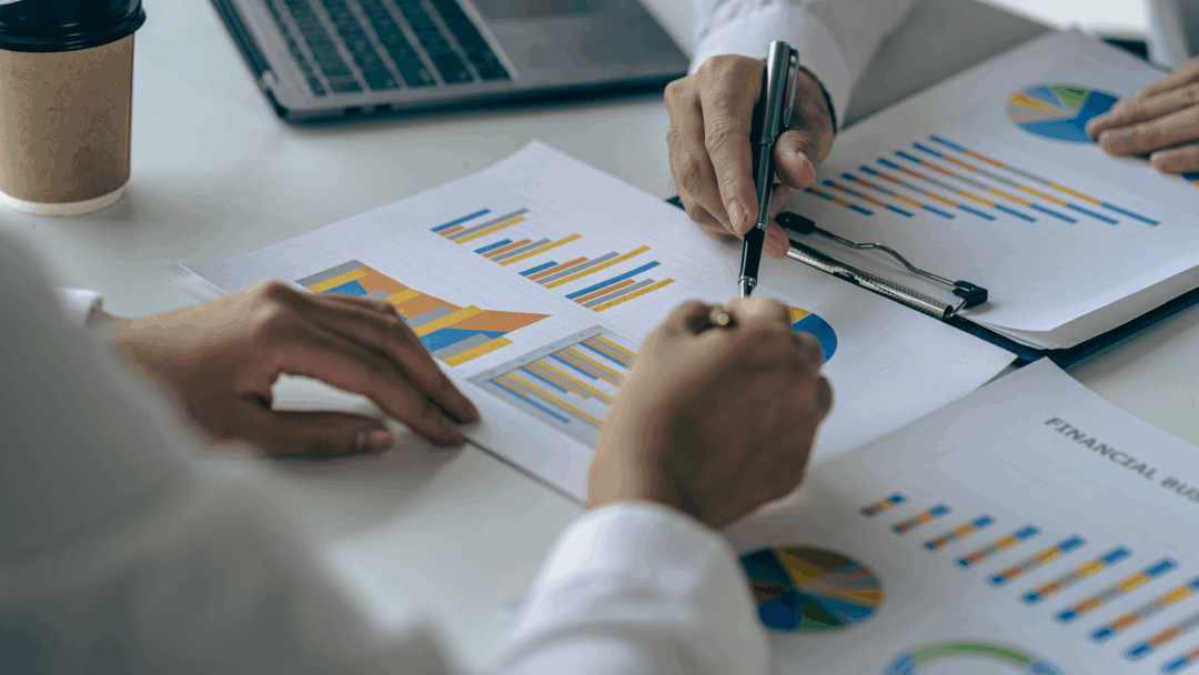 Two people reviewing printed charts and graphs at a desk, pointing at data with a pen near a laptop.
