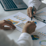 Two people reviewing printed charts and graphs at a desk, pointing at data with a pen near a laptop.