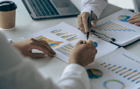 Two people reviewing printed charts and graphs at a desk, pointing at data with a pen near a laptop.