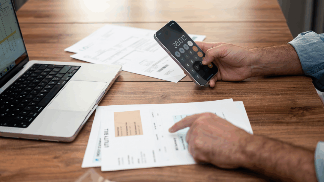 Person reviewing household bills at a table while using a calculator app on their phone next to a laptop.