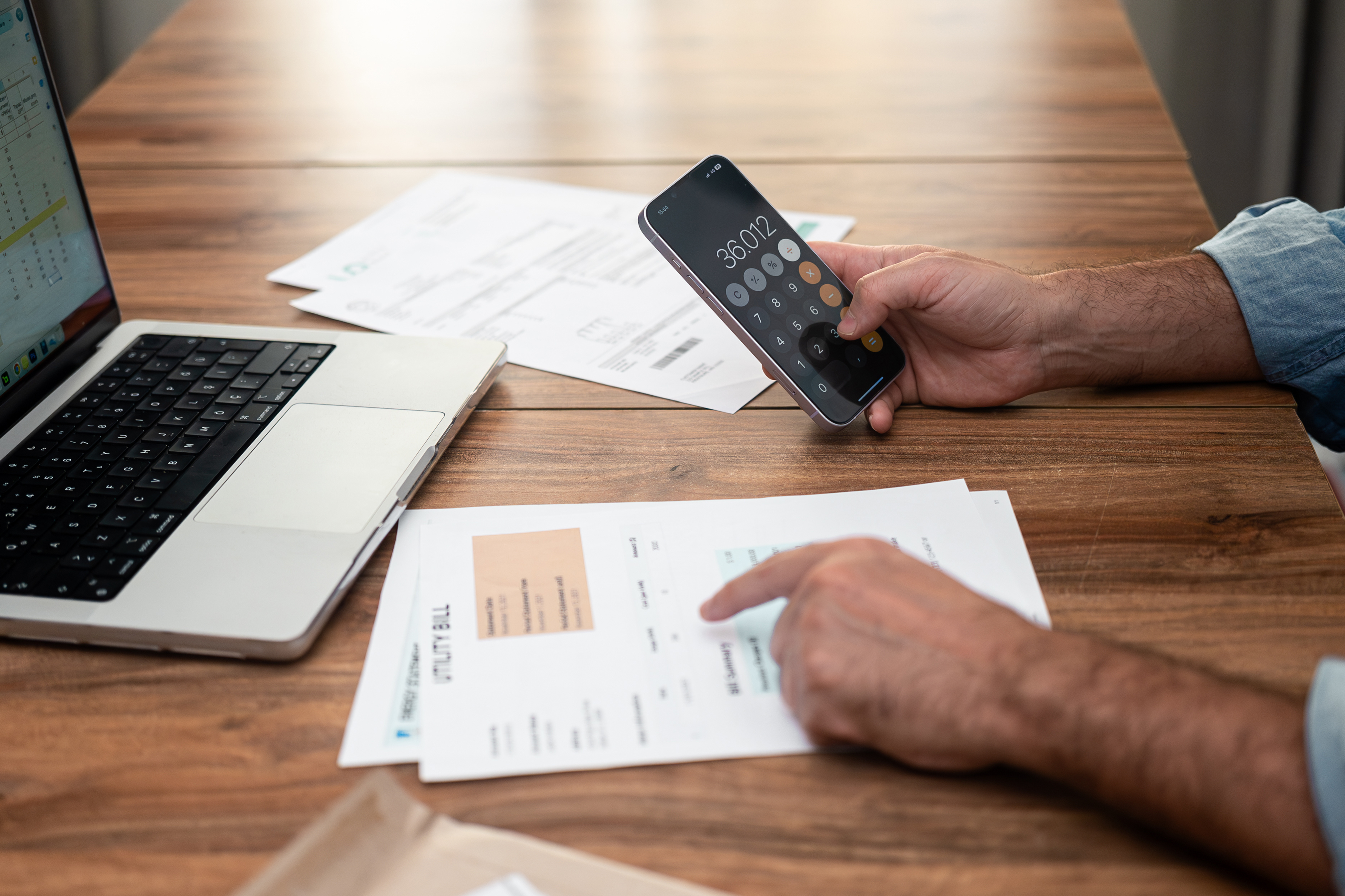 Person reviewing household bills at a table while using a calculator app on their phone next to a laptop.