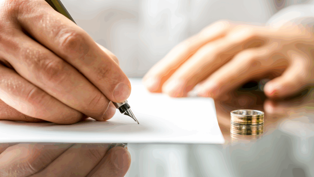 A person signs a document at a desk with a wedding ring placed beside the paper.