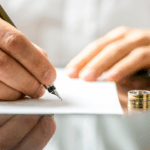 A person signs a document at a desk with a wedding ring placed beside the paper.