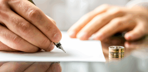A person signs a document at a desk with a wedding ring placed beside the paper.