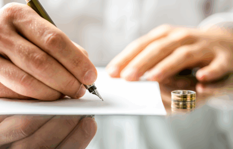 A person signs a document at a desk with a wedding ring placed beside the paper.