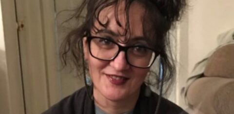 A woman with glasses and dark hair pulled up smiles while holding a bouquet of flowers indoors.