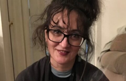 A woman with glasses and dark hair pulled up smiles while holding a bouquet of flowers indoors.