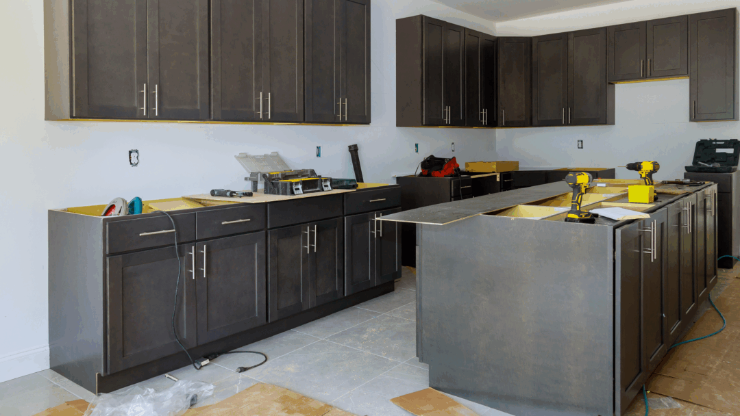 A kitchen under construction with dark cabinets, tools, and materials spread across the floor and counters.