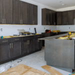 A kitchen under construction with dark cabinets, tools, and materials spread across the floor and counters.