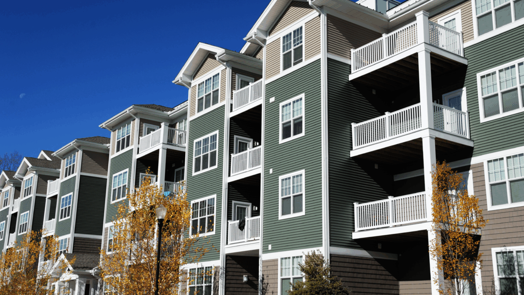 A row of modern apartment buildings with balconies and green siding under a bright blue sky.