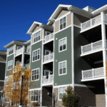 A row of modern apartment buildings with balconies and green siding under a bright blue sky.