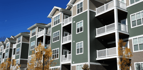 A row of modern apartment buildings with balconies and green siding under a bright blue sky.