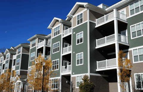 A row of modern apartment buildings with balconies and green siding under a bright blue sky.