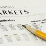 A close-up of financial tables in a newspaper with a pencil pointing toward stock market data.
