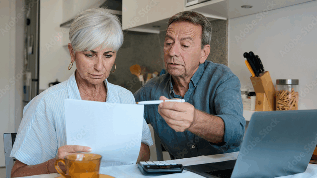An older couple sits at a kitchen table reviewing a document together, with a laptop and calculator nearby.