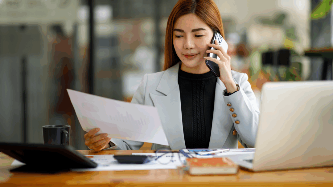 A woman sits at a desk reviewing a document while talking on the phone.