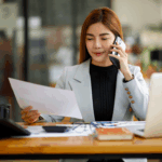 A woman sits at a desk reviewing a document while talking on the phone.