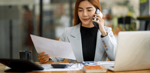 A woman sits at a desk reviewing a document while talking on the phone.
