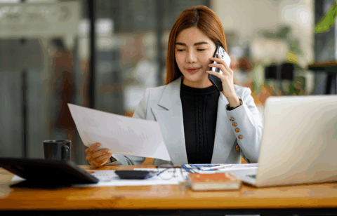 A woman sits at a desk reviewing a document while talking on the phone.